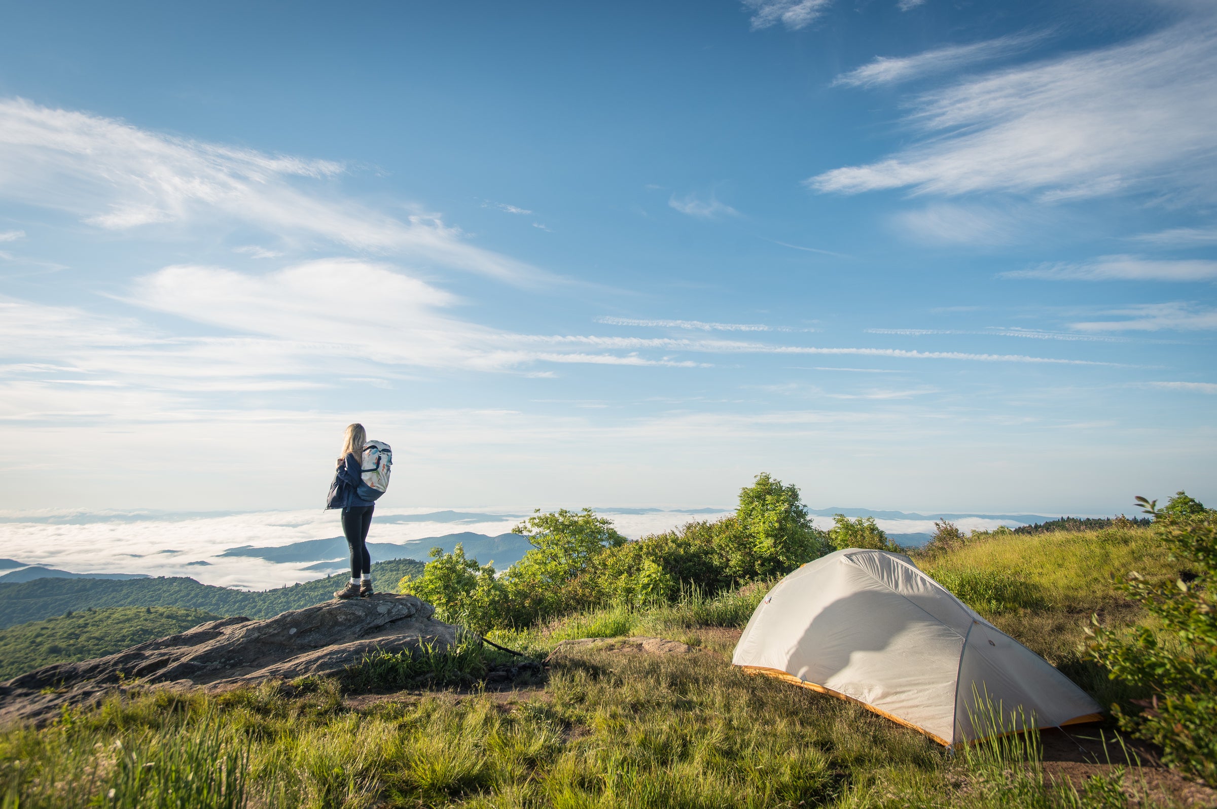 A hiker stands on a rocky outcrop overlooking a scenic mountain landscape, with a tent pitched on grassy terrain nearby under a bright blue sky with scattered clouds.