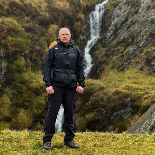 A hiker standing in front of a scenic waterfall in a mountainous landscape, wearing a black waterproof jacket and outdoor gear. The image captures the rugged terrain and natural beauty of the hiking trail.