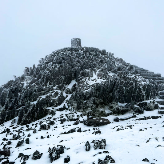 Snowdon summit enveloped in mist, with the iconic marker stone atop frost-covered rocks and a blanket of snow underfoot, capturing the quiet solitude of a winter ascent.