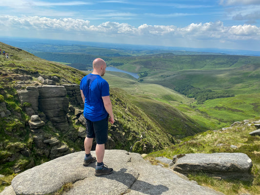 A bald man in a blue shirt and black shorts stands on a rocky overlook, gazing at a lush, green valley below under a clear blue sky.