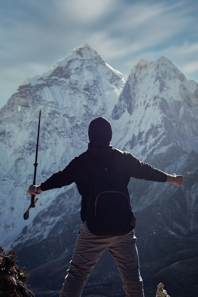 HikeWare - Adventurer with a backpack and trekking pole standing in triumph against a backdrop of majestic snow-covered mountains under a clear sky.