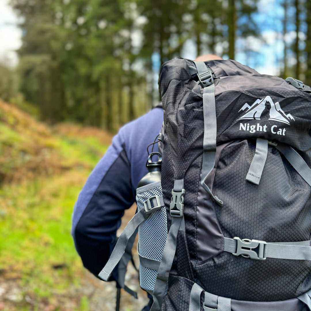 HikeWare - A person from behind wearing a maroon baseball cap with a white logo and a brown tactical backpack with an American flag patch, overlooking a serene blue ocean under a clear sky. Hiking Backpack