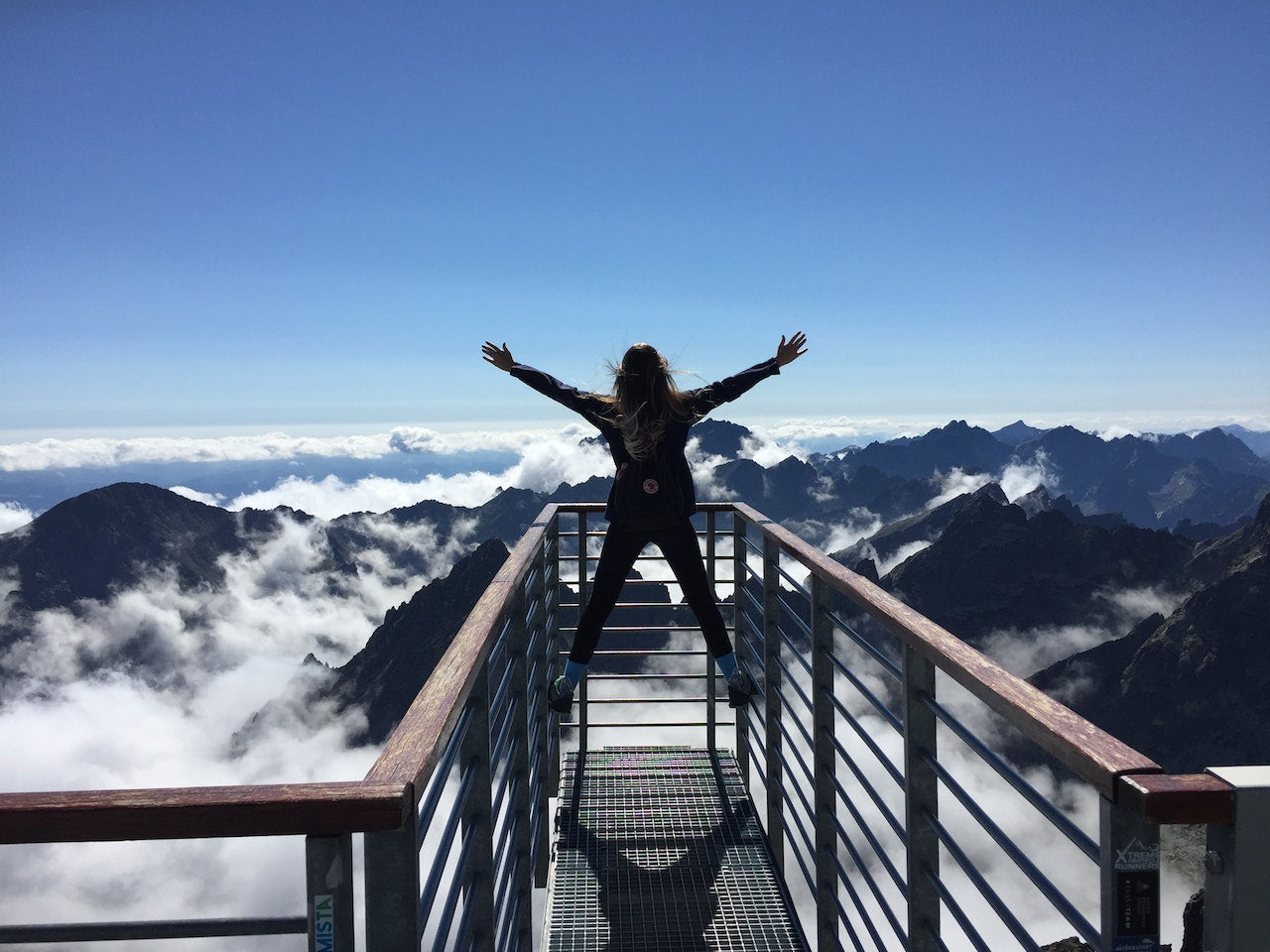 Person with outstretched arms standing on a mountain observation deck above the clouds, celebrating the breathtaking view and sense of achievement